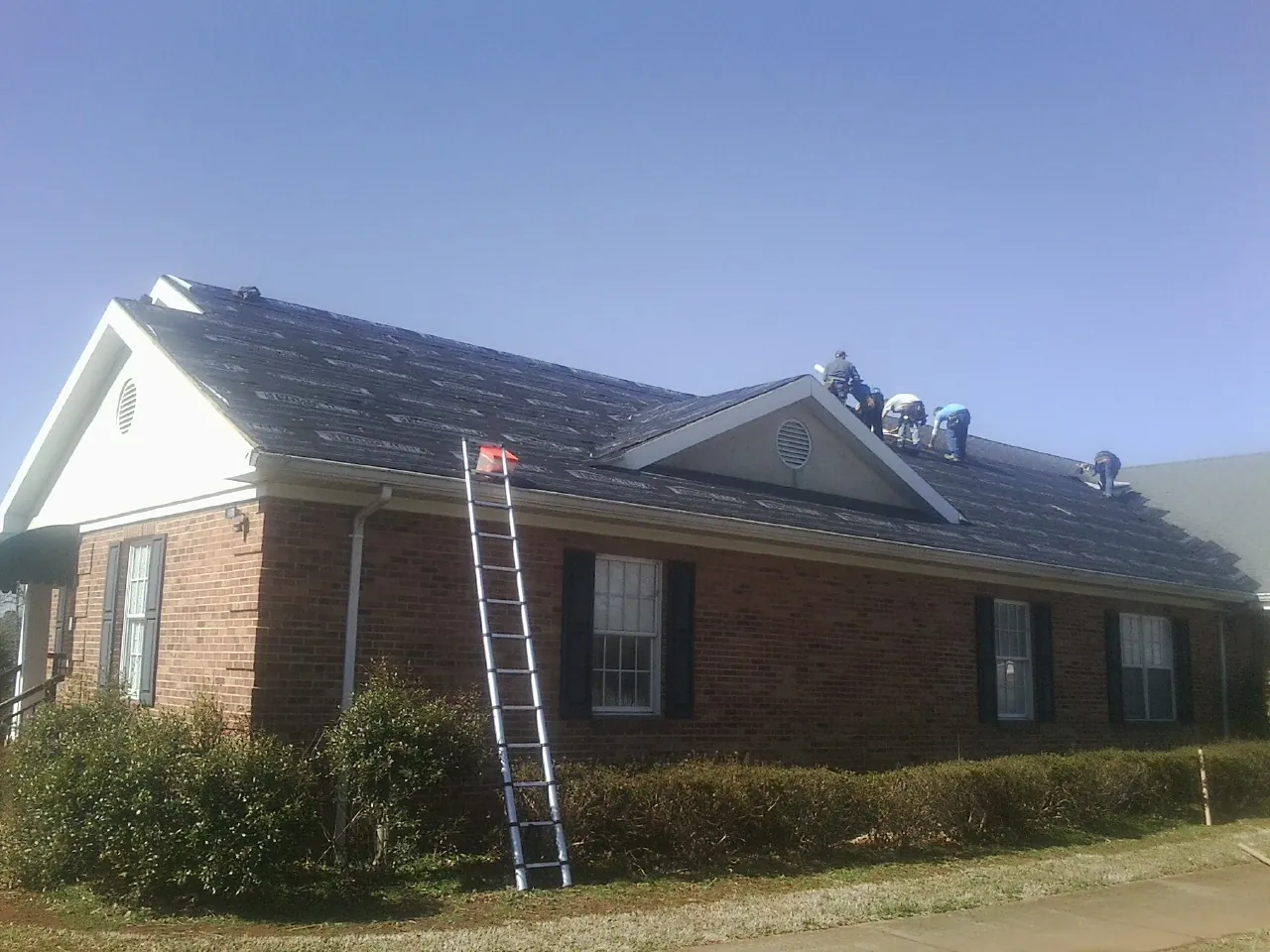 Crew tearing shingles off a brick ranch roof with a ladder set — Emergency Roof Repair work by our crew in Riga